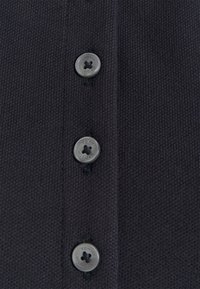 Polo shirt en tricot noir avec une rangée de trois boutons ronds gris gravés "Tommy Hilfiger". Le tissu présente un tissage fin et texturé.