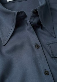 Camicia da uomo blu scuro realizzata in un tessuto liscio e strutturato. Presenta un colletto a punta, bottoni neri e una tasca sul petto.