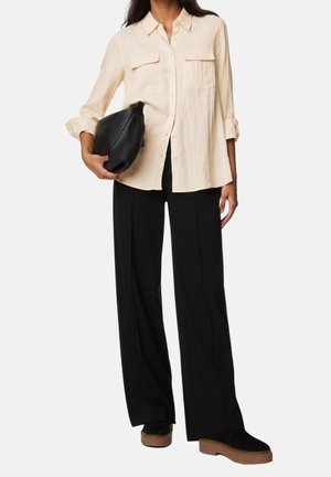 Lichtbeige overhemd met knopen en twee borstzakken, gecombineerd met losse zwarte broeken en zwarte schoenen, terwijl hij een zwarte clutch bag vasthoudt.