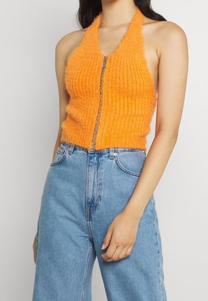 Woman wearing sleeveless orange fuzzy halter top with front zipper and high-waisted light blue denim jeans, standing against plain background.