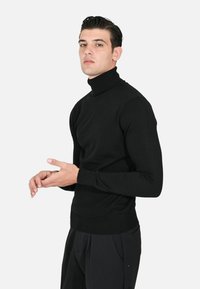 Maglione a collo alto nero realizzato in un tessuto a maglia morbido, caratterizzato da maniche lunghe e un design aderente, indossato con pantaloni scuri.