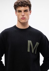 Suéter negro de punto con cuello redondo acanalado. Presenta un gran logo verde "M" en el lado izquierdo del pecho. Textura suave y corte relajado.