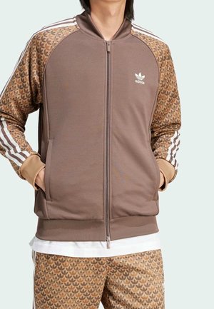 Person trägt eine braune Adidas Trainingsjacke mit gemusterten Ärmeln und passenden gemusterten Hosen, Hände in den Jackentaschen.