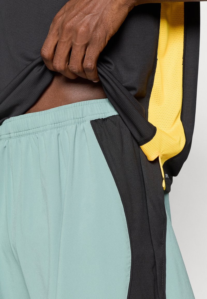 Conjunto deportivo masculino que incluye una camiseta negra texturada y pantalones cortos verde menta claros con acentos negros y un detalle amarillo en el costado.