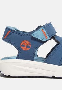 Sininen sandali, jossa on säädettävä hihna, valkoinen pohja, oranssi logo ja kontrastiväriset siniset yksityiskohdat. Teksturoitu päällinen ja pehmeä sisäpohja.