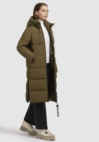 Žena v dlhom olivovo zelenom prešívanom kabáte s kapucňou, bielom roláku, čiernych nohaviciach a béžových robustných čižmách, stojí pred jednofarebným pozadím.