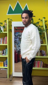 Hvid kabelstrikket sweater med høj hals, kombineret med sorte læderbukser. Baggrunden har en gul væg, grønne dekorative planter og hylder.