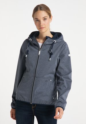 Junge Frau, die eine blaue Kapuzenjacke mit Reißverschluss, Kordeln und Vordertaschen trägt, steht vor einem einfarbigen weißen Hintergrund.