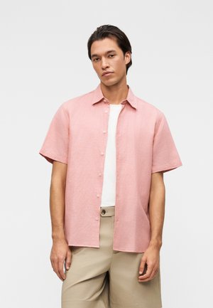 Mlad moški, ki nosi svetlo roza srajco s kratkimi rokavi preko bele majice in bež kratke hlače, stoji pred enobarvnim ozadjem.