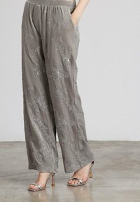 Pantalones grises holgados con bordado floral y lentejuelas, combinados con sandalias de tacón plateadas sobre un suelo de hormigón.
