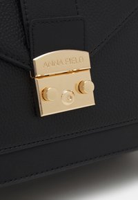 Sac en cuir noir texturé avec une fermoir rectangulaire doré gravé "ANNA FIELD", comportant deux boutons ronds et un mécanisme de verrouillage central.