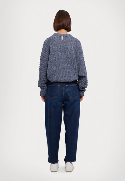 Suéter azul de punto con patrón texturizado y dobladillo elástico, combinado con jeans azul oscuro, visto desde la parte de atrás sobre un fondo neutro.