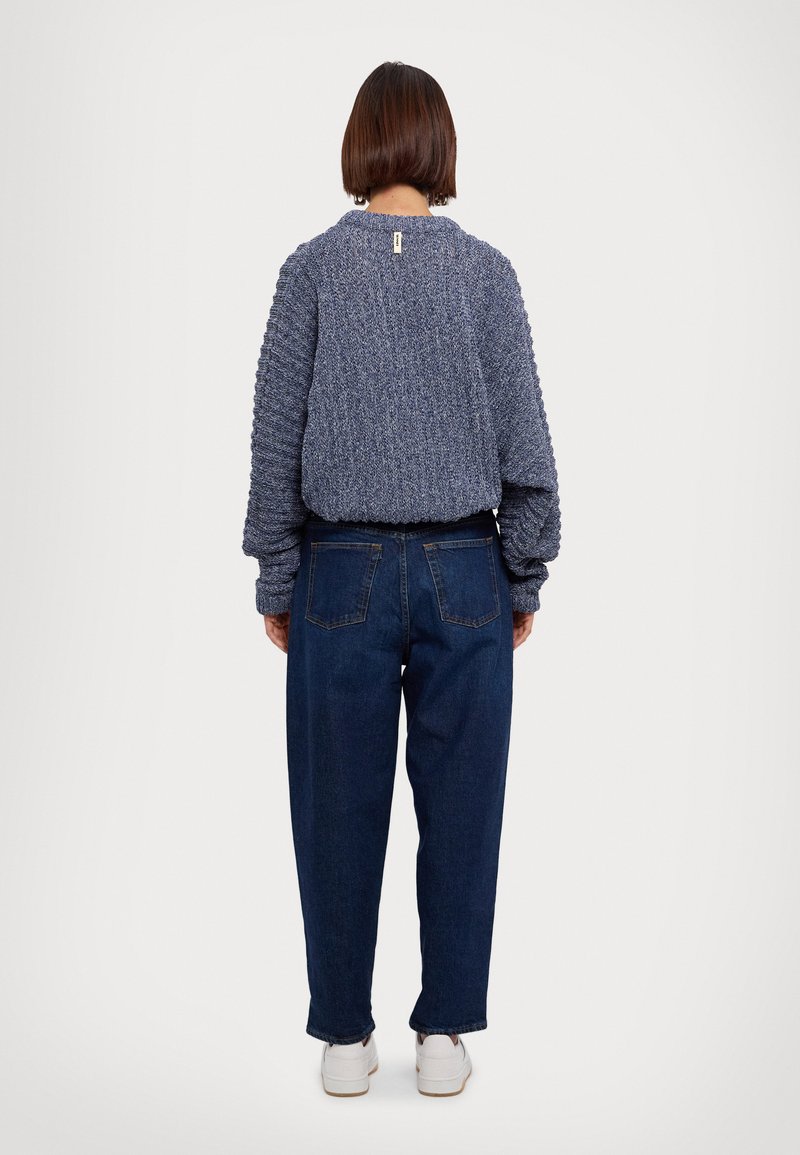 Gebreide blauwe trui met een gestructureerd patroon en elastische zoom, gecombineerd met donkerblauwe jeans, van achteren getoond op een neutrale achtergrond.