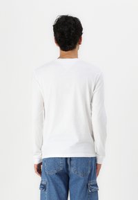 Chemise blanche en coton à manches longues avec un col rond, présentant une texture subtile et un ourlet droit. Portée avec un jean cargo bleu.