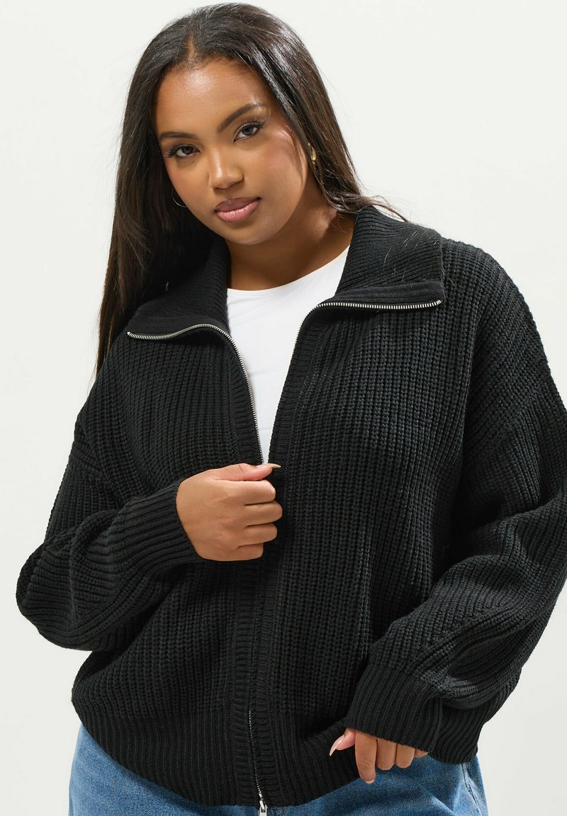 Svart stickad zip-up kofta med hög krage, ribbad textur och oversized passform. Framsidans fickor och långa ärmar med ribbade mudd.