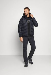 Black padded jacket with a zip, featuring horizontal stitching, worn over a black hoodie and paired with dark trousers and sneakers.