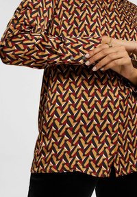 Chemise à motifs dans des tons de marron, moutarde, rouge et noir avec un design géométrique ; elle possède un poignet à bouton et une texture de tissu lisse.