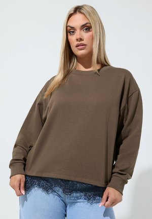 Kobieta o długich blond włosach nosi brązową bluzkę z długim rękawem na czarnym koronkowym detalu i jasnoniebieskie dżinsy na jednolitym tle.