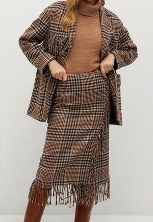 Femme ajuste une jupe à carreaux marron avec franges et une veste assortie portée sur un pull à col roulé couleur chameau avec des bottes marron.