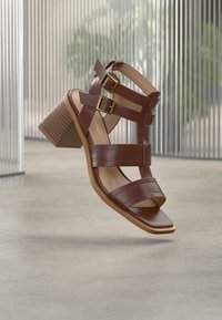 Καφέ σανδάλια με τακούνι, δύο αγκράφες, τετράγωνη μύτη και υφασμένο τετράγωνο τακούνι. Λείο δερμάτινο πάνω μέρος και επίπεδη σόλα.