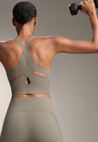 Olivno zelena športna modrček s križnimi naramnicami in izrezanimi detajli, v kombinaciji z visokim pasom usklajenimi legginsami, ki vključujejo subtilno blagovno znamko.