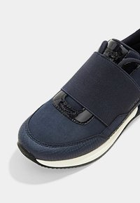 Chaussure de sport bleu marine avec une tige en tissu texturé, une bande élastique et un accent lisse en cuir verni. Dispose d'une semelle en caoutchouc blanche et d'un design à bout rond.
