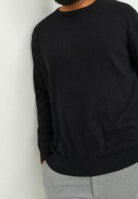 Homme portant un pull noir à manches longues et un pantalon gris clair, debout avec une main dans la poche contre un mur clair uni.