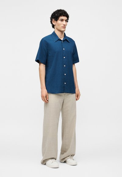 Άνδρας που φορά μπλε πουκάμισο με κοντά μανίκια και κουμπιά, μπεζ παντελόνι φαρδιάς γραμμής και λευκά αθλητικά παπούτσια σε απλό φόντο.
