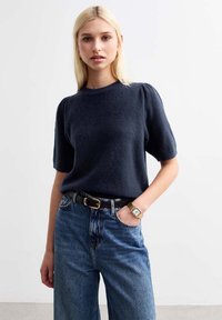 Maglione navy a maglia con maniche corte a palloncino, abbinato a jeans in denim blu a vita alta e un orologio dorato, caratterizzato da un tessuto testurizzato.