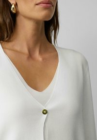 Witte cardigan met een V-hals en een enkele groene knoopsluiting. Zachte gebreide textuur, gelaagd over een bijpassend witte top. Gouden druppeloorbel zichtbaar.