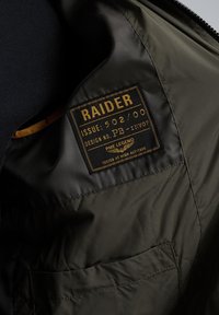Wnętrze oliwkowej kurtki z pikowaną teksturą, z brązową metką z napisem "RAIDER" oraz detalami wzorniczymi i kieszenią.