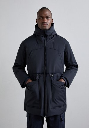 Must pähe ja kõrge kraega must isoleeritud parka. Omab kaht esi-taskut ja täispikkuse lukku, valmistatud säravast ja vastupidavast kangast.