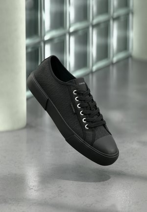 Zwarte laag model sneaker met een gestructureerd patroon, zwarte zool en veters, zwevend boven een gepolijste betonnen vloer bij glasblokken.