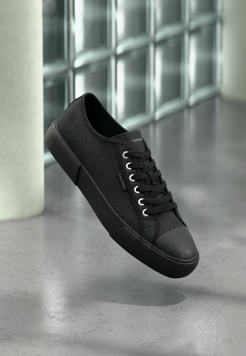 Baskets basses noires avec motif texturé, semelle et lacets noirs, flottant au-dessus d'un sol en béton poli près de blocs de verre.