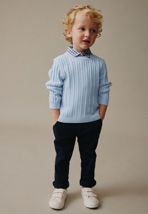 Jong kind met krullend blond haar, gekleed in een blauwe kabelgebreide trui, geruit overhemd, donkere broek en witte sneakers, staand met handen in de zakken.