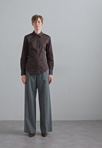 Camicia marrone a maniche lunghe con colletto, abbinata a pantaloni grigi a gamba larga. Entrambi i capi presentano texture lisce e vestibilità su misura.