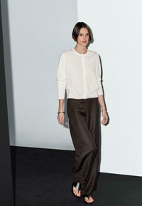 Una donna con un taglio di capelli a caschetto indossa un cardigan chiaro e pantaloni ampi scuri, camminando contro uno sfondo minimale di grigio e nero.