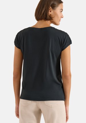 Mujer con cabello corto y castaño lleva una camiseta negra lisa de manga corta y pantalones beige claro, mirando hacia atrás sobre un fondo blanco.