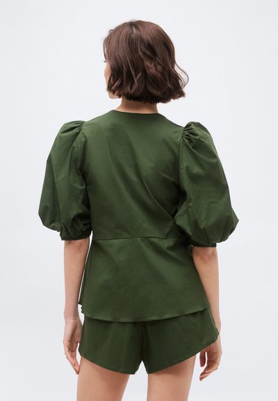 Γυναίκα με κοντά καστανά μαλλιά που φοράει τοπ με φουσκωτά μανίκια σε ελιά πράσινο χρώμα και σορτς που ταιριάζουν, στέκεται γυρισμένη προς τα πίσω με φόντο λευκό.