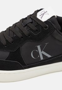 Černé tenisky vyrobené z semišové a syntetické látky; mají výrazné šedé logo "ck", kulaté tkaničky a bílou gumovou podrážku.