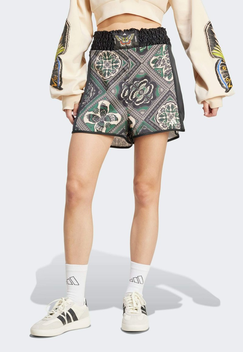 Personne portant un short taille haute à motifs avec une ceinture froncée, détail papillon, chaussettes de sport blanches et baskets, debout les bras détendus.