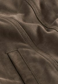 Bruine suede stof met een zachte textuur, met zichtbare stiksels en een subtiele glans. Bevat een klein zakje als detail.