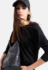 Borsa nera con texture e motivo geometrico, abbinata a una maglietta nera a maniche lunghe. Il modello indossa un berretto abbinato con un design del logo.