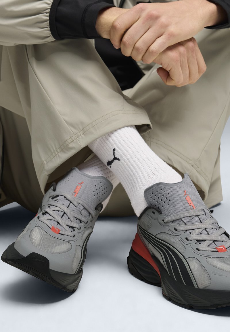 Chaussures de sport grises avec accents rouges, dessus texturé et semelle noire, portées avec des chaussettes blanches côtelées ornées d'un logo noir, sur un fond neutre.