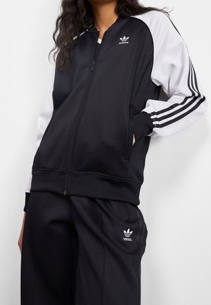 Osoba ubrana w czarny dres Adidas z białymi rękawami i logo Adidas na piersi oraz spodniach, stoi z jedną ręką w kieszeni.