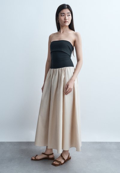 Jeune femme debout, portant un haut noir sans bretelles, une longue jupe beige et des sandales marron, regardant vers sa gauche sur un fond clair uni.