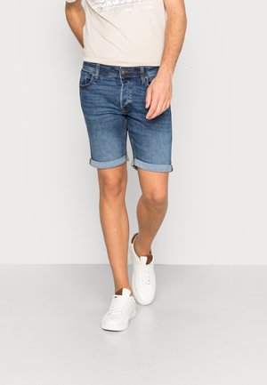 Uomo che cammina indossando pantaloncini di jeans blu risvoltati, sneakers bianche e una t-shirt beige con grafica, davanti a uno sfondo chiaro uniforme.