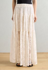 Weißer Maxikleid mit Spitzenmuster, mit einem schichtweisen Design, elastischem Bund und komplizierten Mustern, kombiniert mit schwarzen flachen Sandalen auf einem Holzboden.