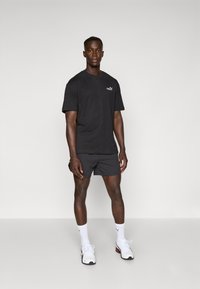 Sort kortærmet T-shirt med et logo på brystet, parret med mørke shorts. Hvide strømper og atletiske sko fuldender outfittet.