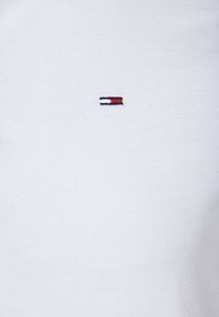 Tissu blanc avec une petite broderie de logo rectangulaire présentant des bandes horizontales noires, blanches et rouges centrées sur la surface.